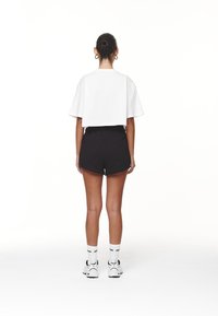 Beskåret hvid t-shirt med korte ærmer, parret med sorte athletic shorts. Modellen har hvide sneakers og sokker med logo på, set fra bagsiden.