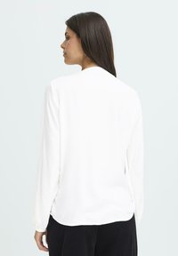 Frau mit langen dunklen Haaren, die eine weiße Bluse mit langen Ärmeln und schwarze Hosen trägt, mit dem Rücken zu einem schlichten hellen Hintergrund gewandt.