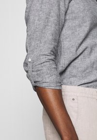 Harmaa pellavapaita, jossa on käännetyt hihat ja napit kalvosimissa. Alhaalla näkyvät beiget housut, sileä rakenne ja rento istuvuus.