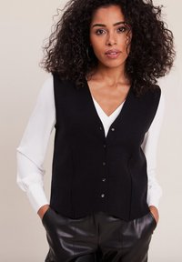 Gilet di lana nera con scollo a V, cinque bottoni e tasche laterali, abbinato a una camicia bianca a maniche lunghe e pantaloni di pelle neri.