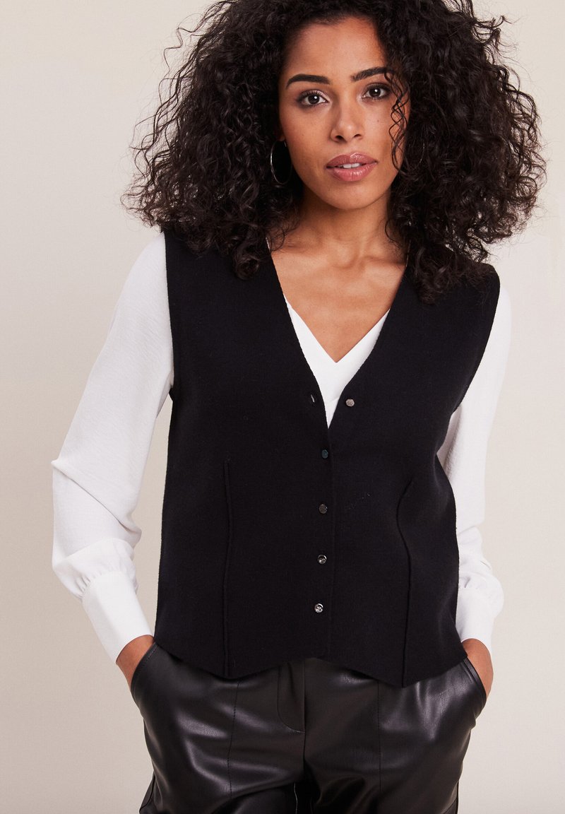 Gilet di lana nera con scollo a V, cinque bottoni e tasche laterali, abbinato a una camicia bianca a maniche lunghe e pantaloni di pelle neri.