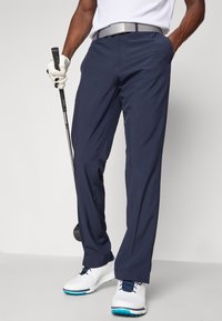Námořnické golfové kalhoty vyrobené z lehké tkaniny, vyznačující se volným střihem, hladkým povrchem a šitými švy. Spárovány s bílými golfovými botami.