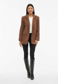 Nainen hymyilee, pukeutuneena ruskeaan mokkanahkatakkia, valkoiseen toppiin, mustiin tiukkoihin farkkuihin ja polvimittaisiin mustiin nahkasaappaisiin, seisten valkoista taustaa vasten.