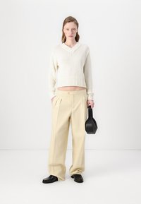 Pulover crem tricotat cu decolteu în V, pantaloni bej plisati, geantă neagră și pantofi negri, stând în fața unui fundal deschis.