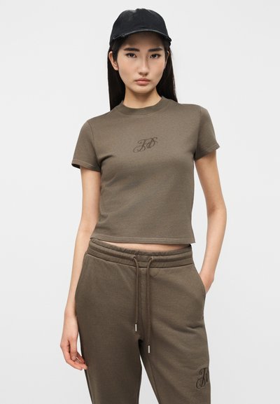 Νεαρή γυναίκα που φοράει ένα καφέ κοντό μπλουζάκι και αντίστοιχο φόρμα παντελόνι, μαύρο καπέλο, στέκεται με το ένα χέρι στην τσέπη μπροστά σε λευκό φόντο.