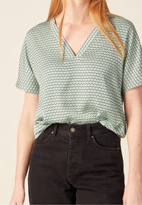 Donna che indossa una blusa a maniche corte con scollo a V a fantasia verde chiaro, infilata in jeans neri a vita alta.