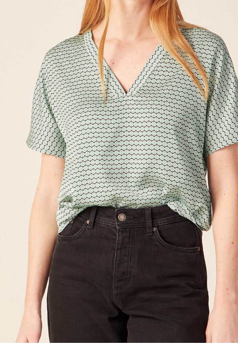 Donna che indossa una blusa a maniche corte con scollo a V a fantasia verde chiaro, infilata in jeans neri a vita alta.