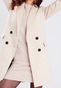 Béžový dvojřadý kabát z vlněné směsi s černými knoflíky, oversized klopami a bočními kapsami, nošený přes světle béžové šaty s žebrovaným lemem.