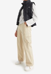 Osoba v béžových cargo kalhotách, bílých teniskách, světle šedé mikině a černé prošívané vestě bez rukávů, stojící s jednou rukou v kapse.