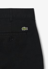 Pantalons noirs avec une texture lisse, dotés d'une poche arrière avec un logo de crocodile brodé en vert et une fermeture à bouton noir.