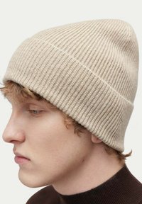 Stickad beige mössa med ribbat mönster, figurnära design och sömlig topp, som bärs på en persons huvud i en lätt vinkel.