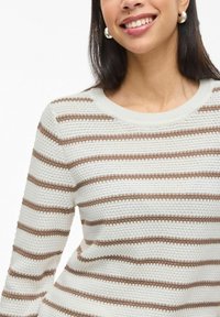 Femme souriante portant un pull tricoté rayé blanc et beige avec un col rond et des boucles d'oreilles argentées en forme d'anneau.