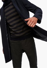 Marineblå frakke med tekstureret stof, båret over en olivengrøn og marineblå mønstret sweater, kombineret med mørke bukser. Har en skræddersyet pasform.