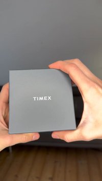 Gray box with a matte finish, featuring the word "TIMEX" in white, centered on the top. Hands holding the box are visible.