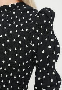 Bluza neagră cu buline albe, având o textură smock, guler ridicat și mâneci bufante cu manșete elastice.