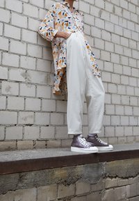 Tryckt skjorta med gula, blå och orange mönster, kombinerad med vita vida byxor och grå höga sneakers, stående mot en tegelvägg.