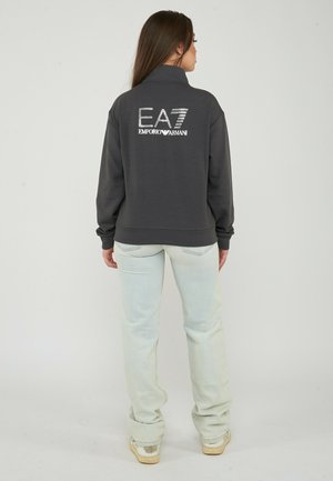Vrouw met lang bruin haar, gekleed in een donker sweatshirt met "EA7 Emporio Armani" op de rug, lichte spijkerbroek en beige sneakers, staand met de rug naar ons toe.
