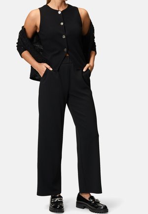 Femme portant un gilet noir sans manches boutonné, un pantalon large noir avec les mains dans les poches, une veste noire tombée des épaules et des mocassins noirs.