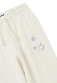 Crème-kleurige sweatpants met een elastische tailleband en zilveren glittersterontwerpen nabij de voork pocket op het rechterbeen.