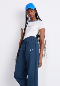 Camiseta blanca con ribete azul y texto "nike", combinada con pantalones deportivos Nike en azul marino. Una gorra azul completa el look atlético.