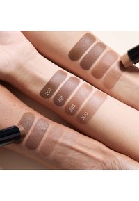 Four arms displaying makeup swatches in various brown shades, labeled 200 to 203. Smooth texture, differing shades range from light to dark.