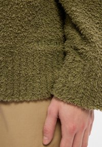 Groene fleece trui met een textuur van pluizig materiaal, voorzien van een geribbelde zoom en 'Husky' branding op de borst. Hand rustend op een taupe stof.