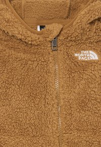 Rjav, puhast flis jakna z zadrgo, ki ima belo logo The North Face in mehko, teksturirano površino.