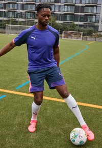 Puma Sportbekleidung: lila kurzärmliges Shirt, marineblaue Shorts mit pinken Akzenten, weiße Socken und pinke Fußballschuhe. Rasenfeld im Hintergrund.