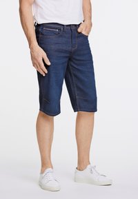 Shorts de mezclilla en azul oscuro, con corte recto, diseño de cinco bolsillos y costuras en contraste, combinados con zapatillas blancas.