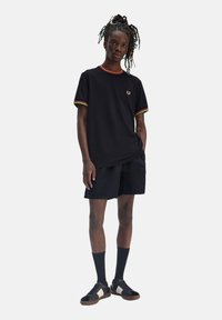 Camiseta negra con ribetes rojos, amarillos y verdes, combinada con pantalones cortos negros. Modelo llevando calcetines oscuros y zapatos negros con acentos blancos.