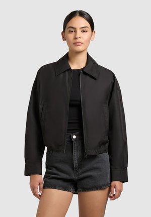 Fiatal, sötét hajú nő, aki fekete cipzáras dzsekit, fekete felsőt és magasderekú fekete farmerrövidnadrágot visel, szürke háttér előtt.