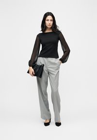 Γυναίκα που φοράει μαύρο ημιδιαφανές τοπ με μακριά μανίκια, γκρι παντελόνι με λεπτές ρίγες, μαύρες γόβες, κρατώντας μαύρη τσάντα, στέκεται μπροστά σε ένα απλό φόντο.