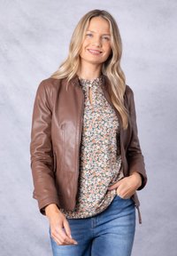 Brown leather jacket with a zip front and side pockets, worn over a floral-patterned blouse. Model has long hair and is smiling.