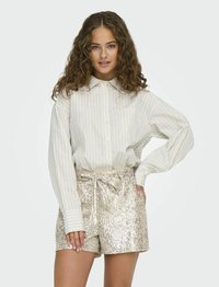 Chemise à rayures à manches longues beige et blanche, associée à un short doré brillant, recouvert de sequins et noué à la taille.