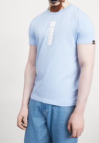 Světle modré tričko s krátkým rukávem z měkké tkaniny s bílým logem "ALPHA". Obsahuje kulatý výstřih a malou černou nášivku na rukávu.