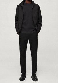 Veste légère noire avec un col montant, portée sur une chemise noire à manches longues assortie et un pantalon noir slim, complétée par des chaussures noires.