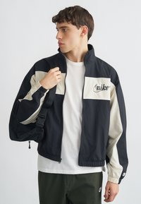 Μαύρο και κρεμ μπουφάν Nike με φερμουάρ μπροστά, ελαστικά μανίκια και λογότυπο. Το μοντέλο κρατάει ένα λουράκι, φοράει λευκό μπλουζάκι και πράσινο παντελόνι.