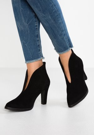 Jambes portant un jean bleu à ourlet effiloché et des bottines noires en daim à talons hauts avec une découpe latérale profonde.