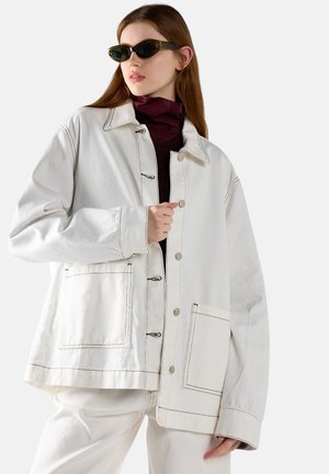 Mujer con chaqueta blanca oversize con grandes bolsillos y gafas de sol oscuras, posando con la mano cerca de los botones y mirando hacia la izquierda.