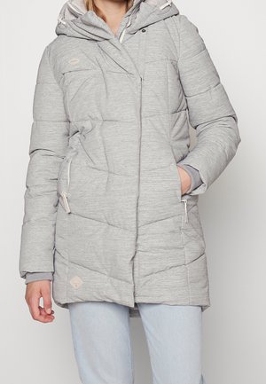 Osoba ubrana w jasnoszarą pikowaną kurtkę zimową z kapturem, kieszeniami na zamek oraz lewą ręką w kieszeni, w połączeniu z jasnoniebieskimi dżinsami.