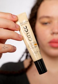 Tubetto di crema CC beige chiaro con tappo nero, etichettato "MÁDARA," presenta "Antinquinamento all'Acido Ialuronico" e "Tinta: Media," materiali riciclabili indicati.