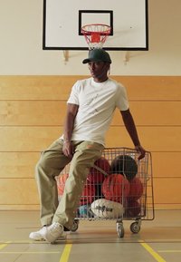 A bevásárlókocsi több sportlabdát tartalmaz. Egyén ül mellette világos ingben, olíva zöld nadrágban és fehér sportcipőben egy edzőteremben.