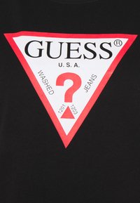 T-shirt nera con logo triangolare bianco e rosso con il testo "GUESS U.S.A." e un punto interrogativo, che fa riferimento a "WASHED" e "JEANS."