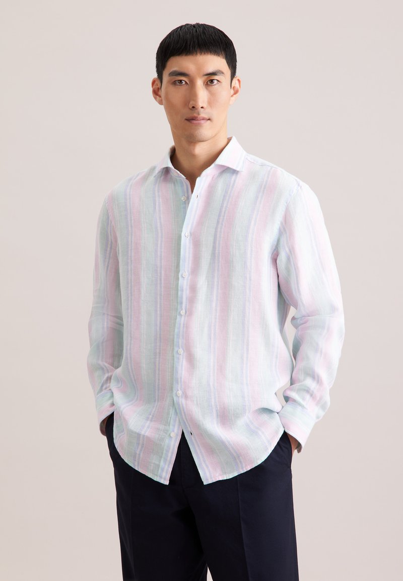 Homme portant une chemise à rayures pastel à manches longues boutonnée, mains dans les poches, sur un fond clair uni.
