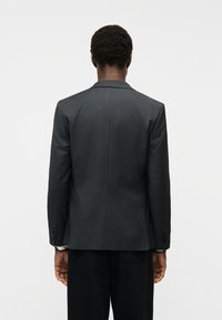 Άνδρας που φαίνεται από πίσω φοράει σκούρο γκρι σακάκι και μαύρο παντελόνι μπροστά σε απλό φωτεινό φόντο.