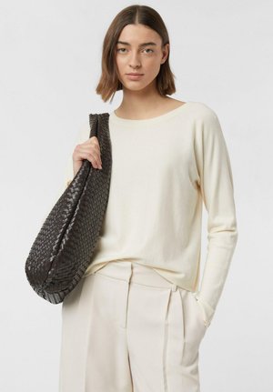 Femme portant un pull et un pantalon couleur crème, tenant un grand sac à bandoulière en cuir tressé brun foncé d'une main.
