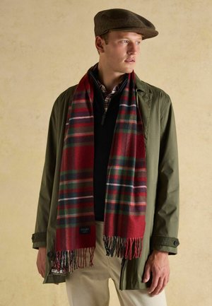 Bărbat tânăr purtând o șapcă maro cu cozoroc plat, jachetă verde măsliniu, pulover negru cu fermoar, cămașă în carouri și fular roșu tartan, pe un fundal simplu bej.