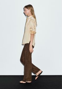 Beige Satin-Hemd mit Knöpfen, hochgekrempelten Ärmeln, kombiniert mit dunkelbraunen Wildlederhosen und schwarzen minimalistischen Sandalen. Profilansicht vor einem schlichten Hintergrund.