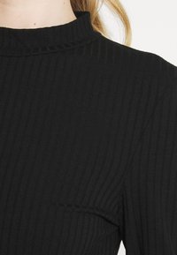 Svart ribbad topp med hög krage, som har ett strukturerat tyg och en figurnära design. Nära vy som framhäver tygmönstret och halsringningen.