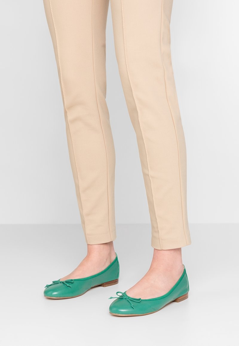Jambes portant un pantalon beige et des ballerines en cuir vert avec petits nœuds, debout sur un sol et un arrière-plan gris clair.